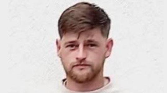 Alex is standing against a white background. He is wearing a white jumper and is looking straight ahead. He has brown hair and a short brown beard.