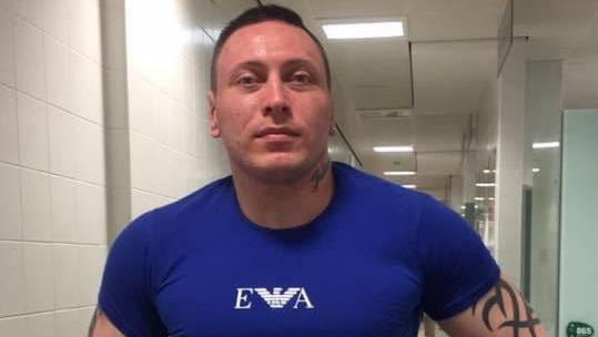 A man with short brown hair and a neck tattoo. He is wearing a dark blue top with white writing that reads 'EA' with a bird emblem. He also has a black tattoo on his upper arm.