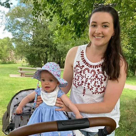 A little baby girl with a colourful spotty hat on and blue eyes beside her mum who has her hand on her. Mum has brown long hair and brown eyes and is wearing a white round necked vest top with maroon on it. They are in a park with a bench and it is sunny.