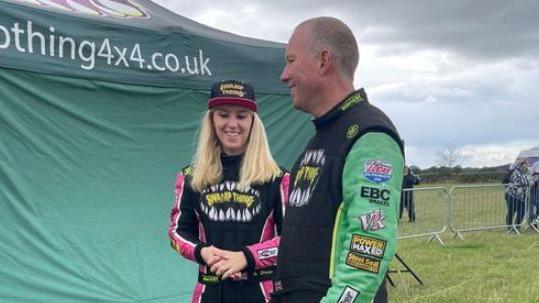 Georgina and Tony stand together smiling outside a tent in a field wearing leathers.
