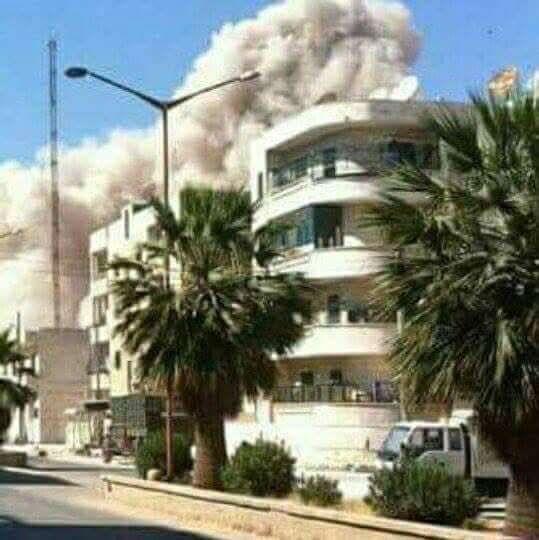 A cloud of smoke and debris rises behind a four-storey apartment block and two palm trees
