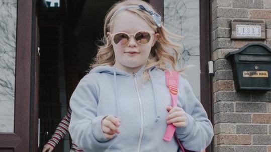 Claudia walking towards the camera wearing a pair of black jeans, a grey zip-up hoodie and white trainers. She has blonde hair, pink sunglasses and a pink backpack. In the background is a house with the front door open.