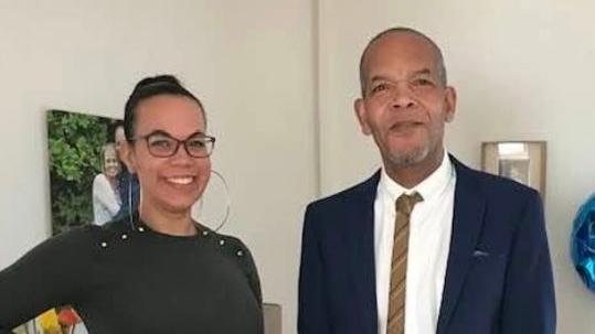 Charlotte Tobierre is wearing a black long-sleeved top, large silver hoop earrings and glasses. Her dad is standing next to her, wearing a navy suit jacket, white shirt and a dark yellow or brown tie.