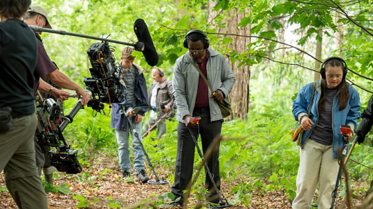 A group of six people stand in woodland, one of them is filming with a large camera, and a boom mic is also visible