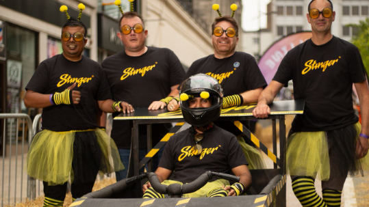 Four team members and their driver stand next to their kart wearing yellow and black T'shirts, socks and netting as skirts
