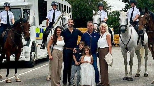 Two men in dark clothing, with two women, both wearing white tops, with their arms around each other. Two children are stood in front of them. Behind are four horses. The two in the middle are white. All of the horses have police riders. They are all smiling into camera.