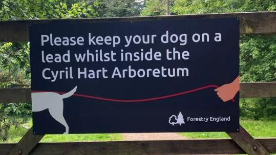 A sign on a fence in a wooded area. The sign reads: "Please keep your dog on a lead whilst inside the Cyril Hart Arboretum".