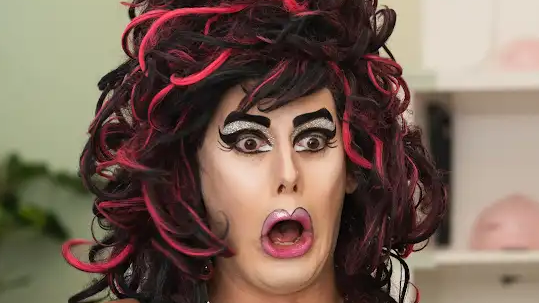A drag queen with pink and black wavy hair, brown eyes, black painted on eyebrows, black eyeliner, silver eyeshadow and red lipstick on. They are pulling a dramatic face and the background behind is blurred.