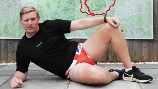 A man wearing a black t-shirt and Union Flag underpants is seen lying in front of a GPS route of a giant penis 