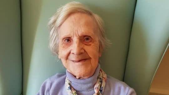 The late Mary ‘Pat’ Thompson smiling and sitting on a green, high-back chair.  She has short, white hair and is wearing a lilac polo-neck jumper and a scarf. 