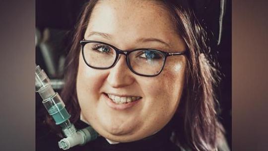 A head shot of a woman looking into the camera and smiling. She has shoulder length brown hair, and is wearing black glasses. She has a medical device, with a tube, coming out of her neck. She is wearing black clothing. 