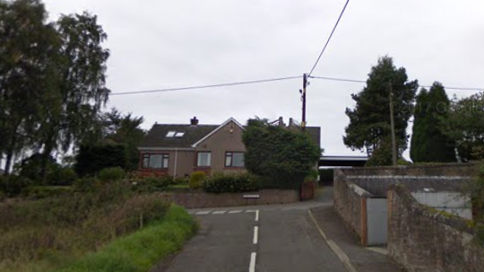 An uphill road leading to a junction with houses on two sides and a field on the left