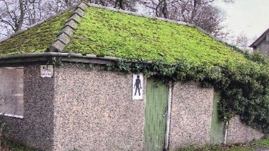 The same pebbledash toilet block back in 2004. It's roof is covered in moss, with vegetation covering its right hand side and one of the doors. One window is covered up. The male toilet sign can be seen on the right, in front of a green door.
