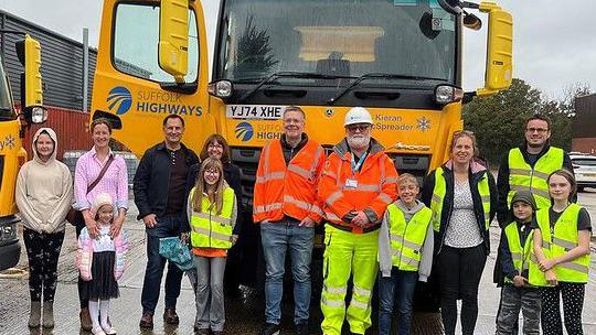 A picture of Suffolk Highways crews standing in front of the Kieran McSpreader gritter with a group of children and their parents. They are standing in a line and smiling at the camera.