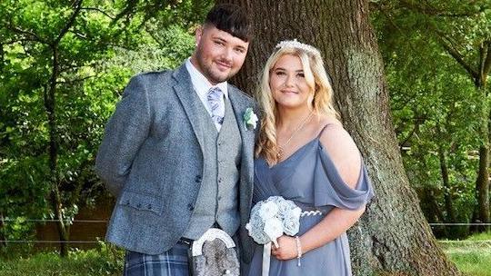 Johnny Baxter stands next to his sister outside in front of a tree, both appearing to be attending a wedding. Mr Baxter has short brown hair and is wearing a grey suit jacket, a grey waistcoat, a purple striped tie and a kilt, while his sister has long blonde hair, a nose piercing and is wearing a grey wrap dress. She is holding a bouquet of white and grey flowers and is wearing a floral head band.