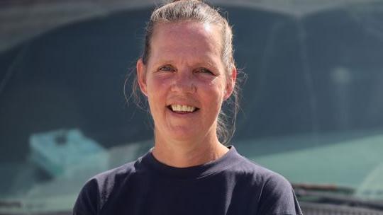 A woman with swept back hair and a blue top is smiling at the camera. Her head and shoulders are visible and the background is indistinct, grey/blue.