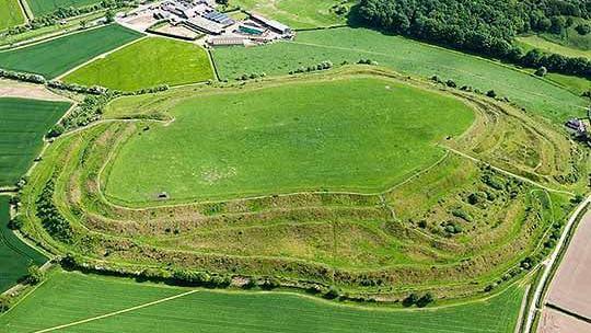 An aerial view of Old Oswestry hillfort. It's a green, grassy plateau with a series of three concentric ridges and slopes leading up to the top. On the left, there's a more complex series of ridges, with three ponds. It's surrounded by farmland and fields - the grass is a bright, vibrant green. 