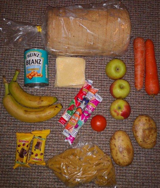 The picture shows a loaf of bread, a bag of pasta, one can of baked beans, some cheese, three apples, two carrots, one tomato, two baked potatoes, two bananas, two malt loaf snacks and three snack size tubes of yoghurt.