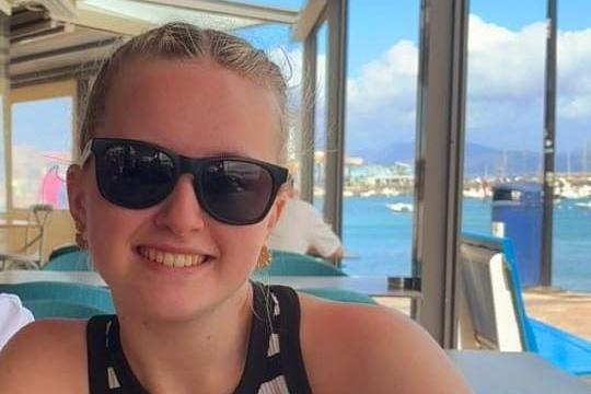 Meg with tied-back blonde hair, wearing sunglasses and a black and white striped vest top. She is sat down, smiling at the camera, with a marina behind her with blue water and white boats.