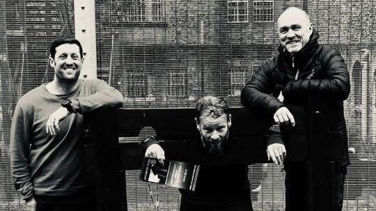 Black and white photo of Chris, Jay and Danny at Shepton Mallet Prison. All three are laughing while Jay has his head and hands through a set of stocks.