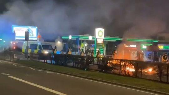 A blurry image of riot police on a road with a petrol station in the background. A fire is lit in front of the police on the road. The view of the road is partially obscured by a hedge.