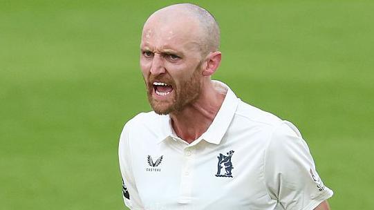 Olly Hannon-Dalby pumps both fists by his side in celebration after taking a wicket for Warwickshire. He has a shaved head and closely clipped bears and is shouting. 