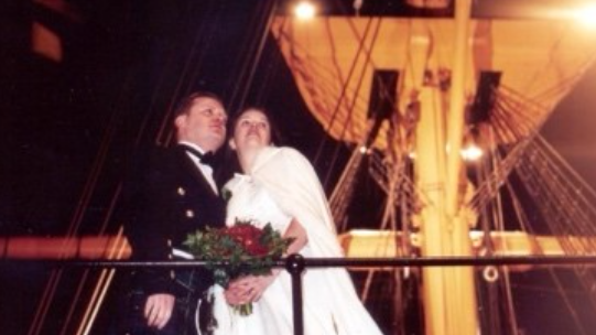 Chris Egan wearing a dark suit with his wife, wearing a wedding dress on a boat. Chris has short dark hair and wears what looks like the tartan of a kilt. Behind them, the mast of the ship is lit up yellow by lights.