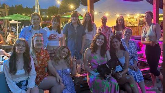 A group of young women wearing colourful outfits sitting in the outside area of a bar with fairy lights strung above them. They are all smiling at the camera. 