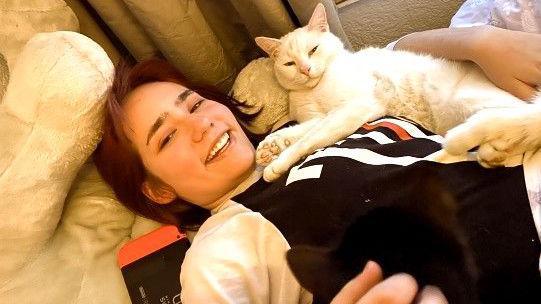Elise Sebastian lies on cushions with curtains in the background, she is smiling and looking at the camera and has a white cat on her lap. She has her arm around the cat. Elise has brown hair and is wearing and black and white top
