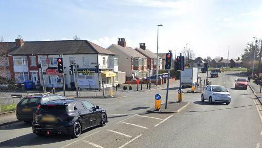 Google streetview of the pedestraian crossing with cars at the traffic lights, and shops and houses in the background