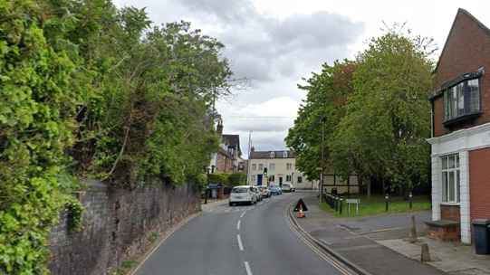 A street going uphill with cars sitting at red traffic lights. To the left is a wall with greenery over the top of it.