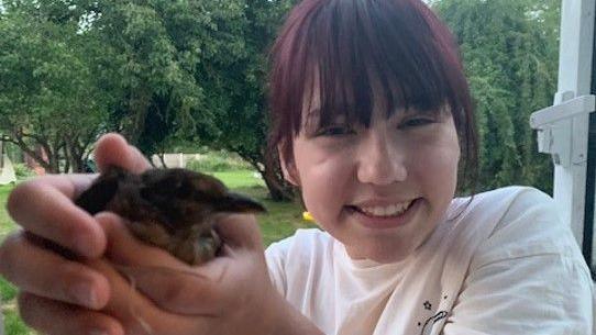 Elise Sebastian, wearing a white t-shirt. She has dark hair which has been pulled back, leaving a long fringe which hangs over her forehead and eyes. She is smiling at the camera, and she is holding a small bird in her hands. There are trees in the background.
