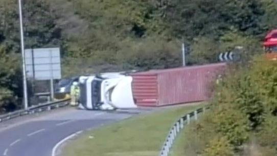 A traffic camera image of a large lorry that has overturned and is on its side in the middle of a road.