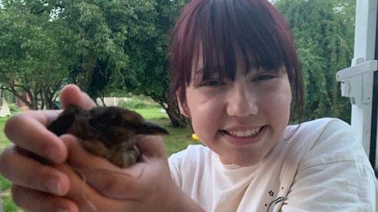Elise Sebastian holds a small bird in her hands. She is smiling happily and is wearing a white T-shirt. Elise has dark brown hair with a fringe which is dyed red. She is outside with trees in .