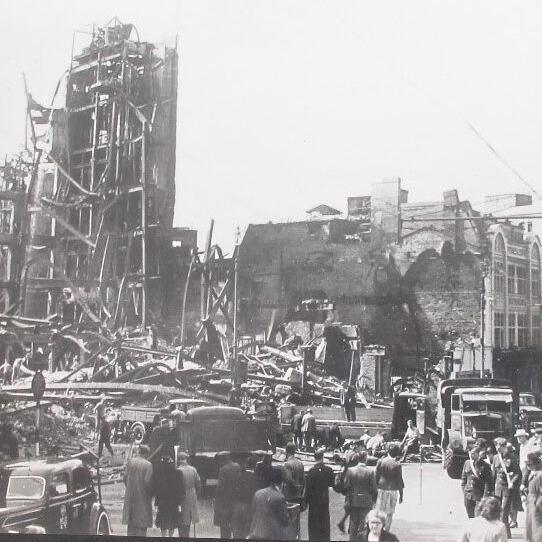 A black and white image showing the shop demolished after bombing during World War Two. Passers by and  vehicles can be seen in the foreground.