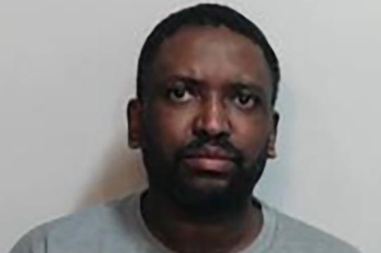 Isaacs has a black moustache and short black hair. He is wearing a grey t-shirt.