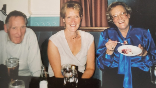 Mr Gwilt is sat on the left with grey hair and wearing a white shirt and trousers. His niece Carole is in the middle, with short blonde hair and wearing a white top. Mr Gwilt's wife Barbara is on the right in a blue shirt and dark skirt. They are smiling at the camera and look to be sat in a pub with drinks on the table in front of them. 