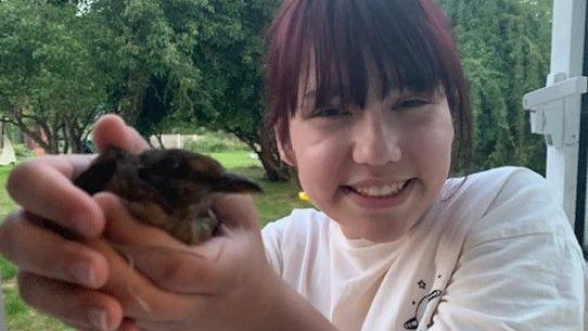 A young girl holding a small chick and smiling at the camera. She is wearing a white top and has fringe over her face