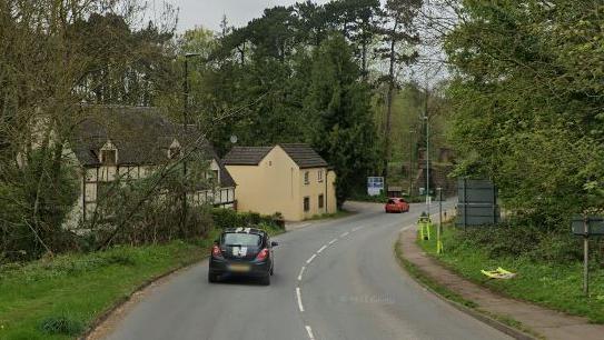 A black Corsa with white stripes drives down a winding B road, with cream coloured and Tudor-style cottages to the left hand side of the road, as a red car drives in front of it. The road is lined with tall trees on a cloudy day.