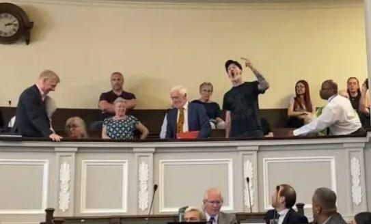 A video still of members of the public sat in a gallery above councillors in the city council chamber. One man, dressed in a black T-shirt and a black hat, is stood up and gesturing with a raised arm towards the councillors.