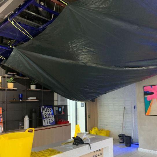 A large black tarpaulin hangs from a ceiling in a building lobby, with buckets placed on the floor to catch water.