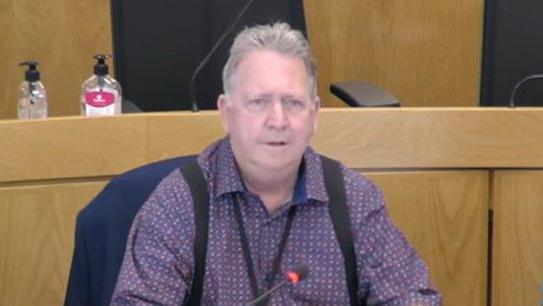 A man with grey hair in a purple patterned shirt and black braces over his shoulders. He appears to be speaking in a council chamber.