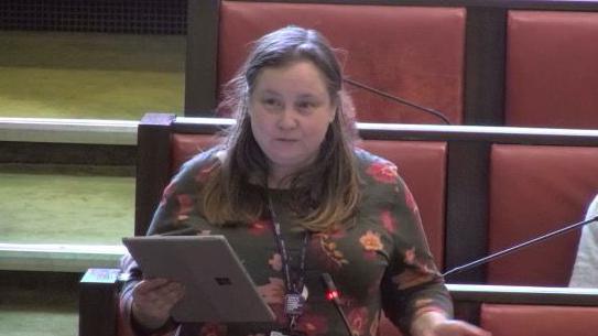 Councillor Sarah Feeney, Labour opposition leader, stands during the 2025 Warwickshire County Council budget setting meeting. She is wearing a flowery top and holding a tablet computer. There are red leather and wooden benches behind her.