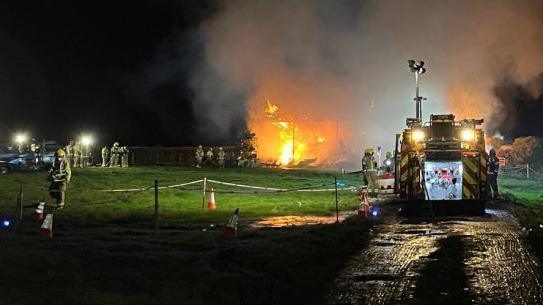 Firefighters in yellow uniforms stand near a blazing barn. One fire engine can be seen.