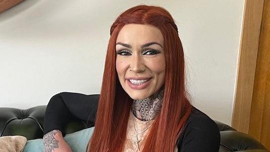 Michelle Moffatt has long ginger hair and smiles at the camera. She has neck and hand tattoos and appears to be sitting on a couch in a living room.