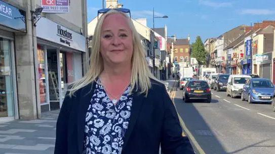 Karen McShane stands in Enniskillen wearing a dark blouse and jacket, she has long blonde hair