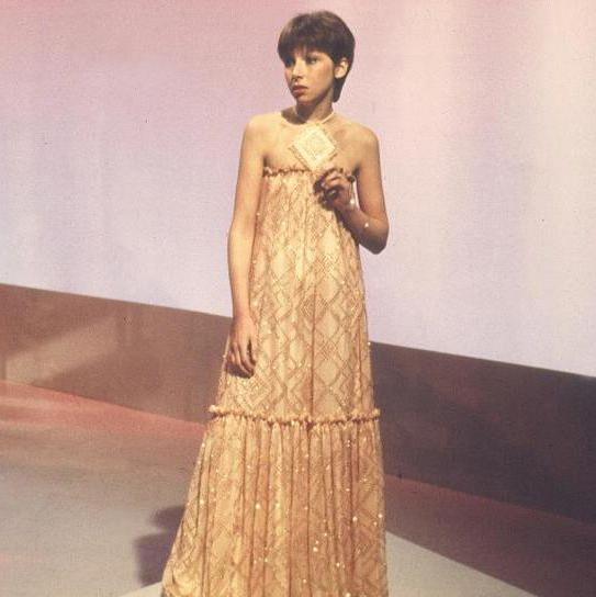 Lena, with short hair, performing in a golden coloured floor-length dress with her bare shoulders visible.