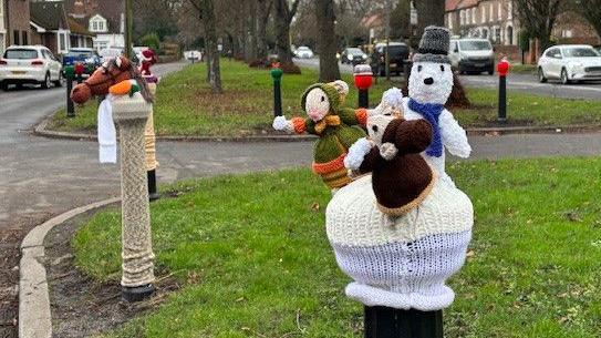 A wide residential street separated by a central grass verge with a line of  well-established trees. Among the knitted characters sitting on bollards are a horse, Father Christmas and three mice characters in Dickensian dress.