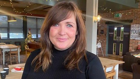 Naomi Chiffi of National Theatre Wales looks into the camera in a large open-plan office, with a poster reading 'Follow Your Dreams' on the wall