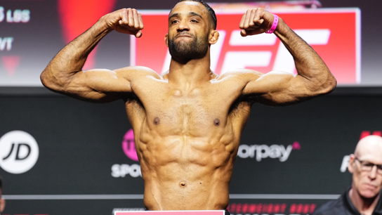A UFC fighter stands with his biceps flexed on a stage. He is topless but wearing black shorts. The background behind him is a black advertising wall with different brands on it. 